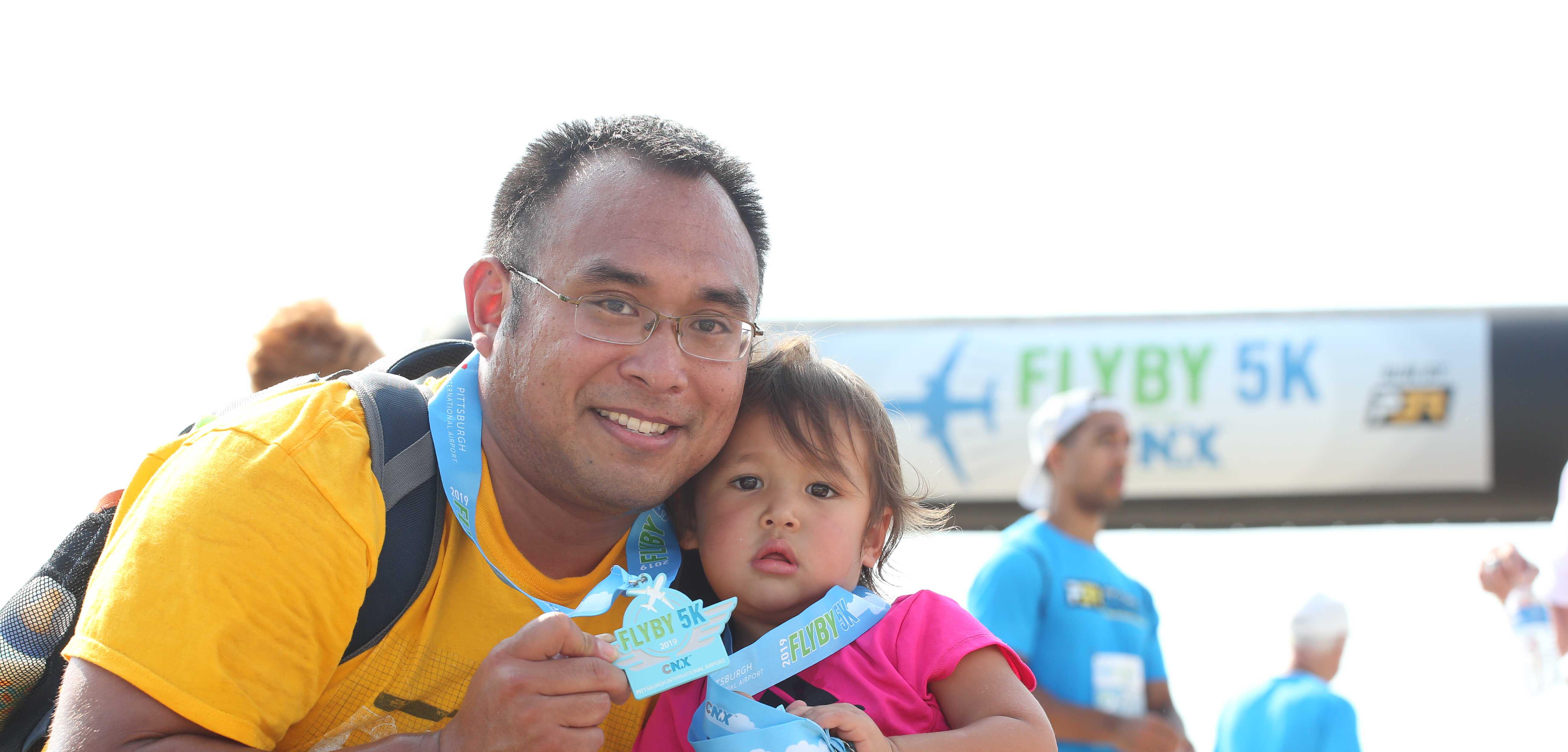 Middle-aged man holding a very young child as they smile with their Fly By 5K medals.