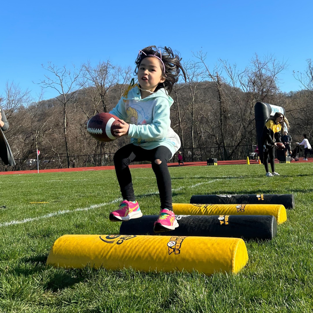 young girl jumping over pads while holding a football