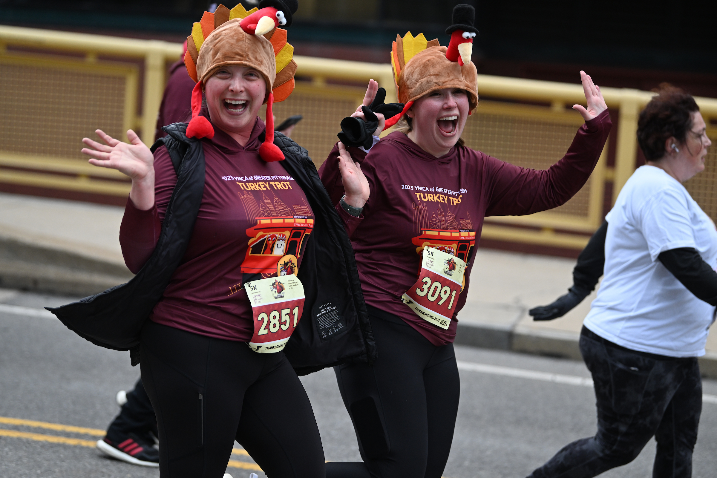 two women wearing turkey hats smiling and waving as they run across the bridge.