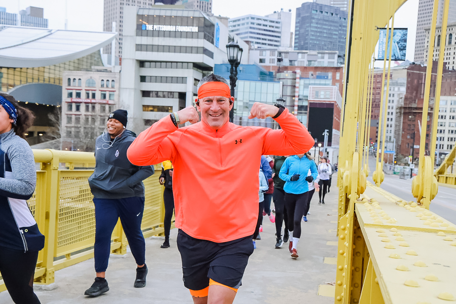 A man running in a vibrant orange shirt flexes while running across the bridge.